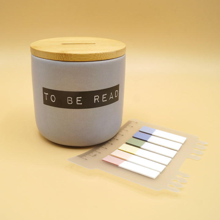 A gray, ceramic jar printed with black lettering reading 'TO BE READ' and a wooden lid. A set of six colored book tabs is next to it.