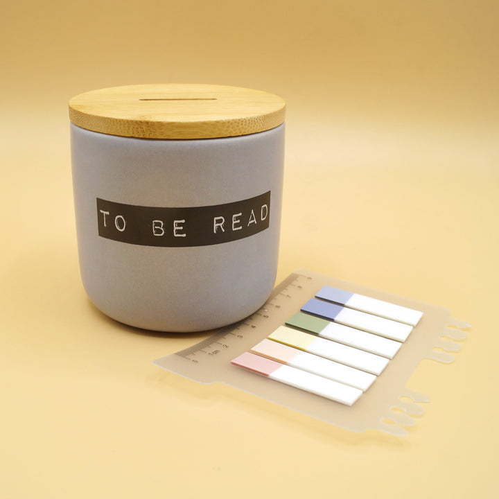 A ceramic "To Be Read" jar with a wooden lid next to colorful bookmark tabs on a beige background.