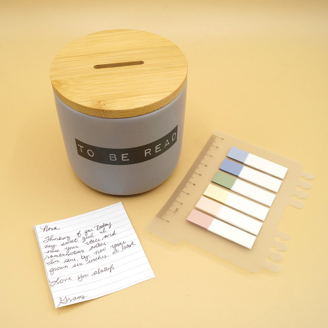 A gray, ceramic jar printed with black lettering reading 'TO BE READ' and a wooden lid. A set of six colored book tabs is next to it along with a piece of paper with handwriting on it