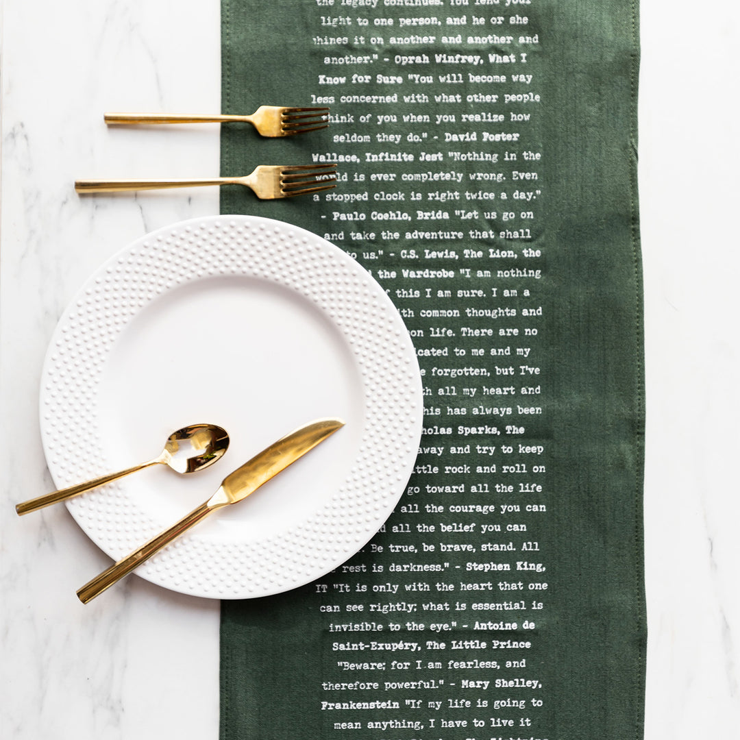 A dark green table runner with quotes from books in white writing down the center is on a marble background. A white plate and gold utensils are on the table runner.