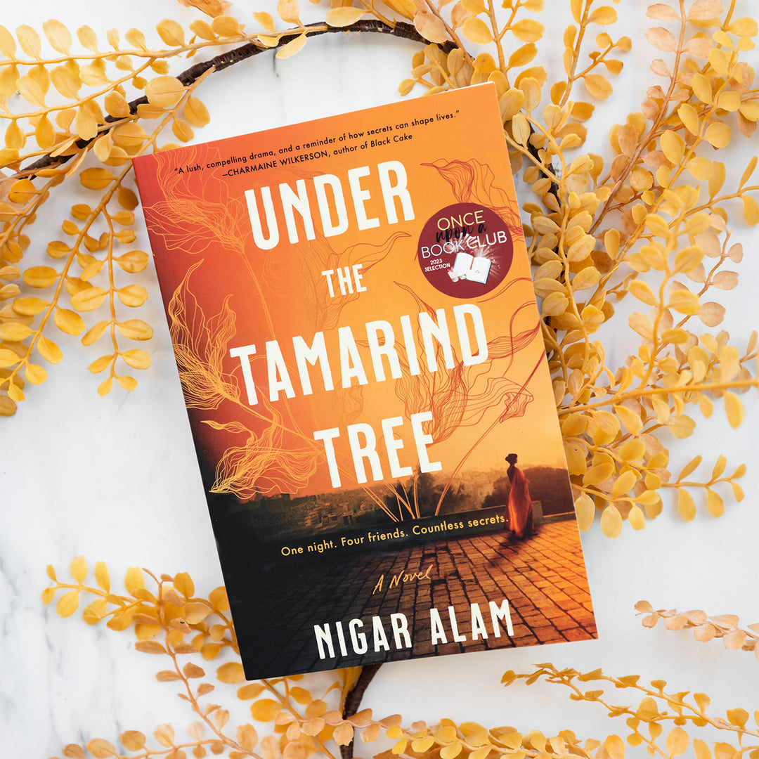 a paperback copy of Under the Tamarind Tree by Nigar Alam sits on yellow/orange leaves