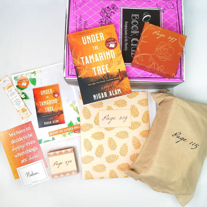 A paperback edition of Under the Tamarind Tree and an orange box sit on a pink Once Upon a Book Club box. In front of the box are a bookmark, quote card, signature card, bookclub kit, beige box, orange and white folder, and beige bag. The boxes and bags all have page numbers.
