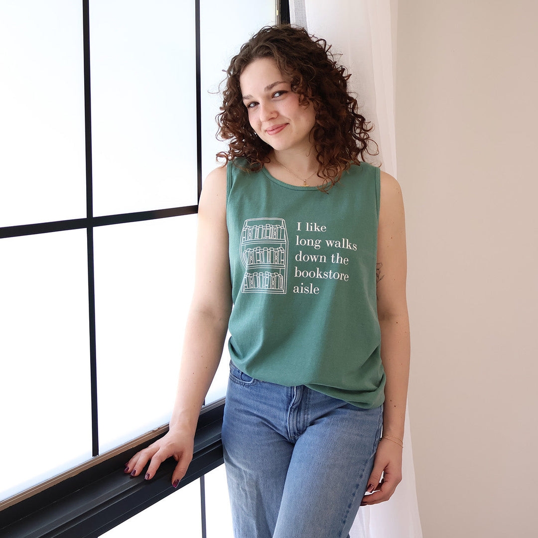 Woman wearing a green tank top with text that reads 'I like long walks down the bookstore aisle' and bookshelf graphic standing by a window.