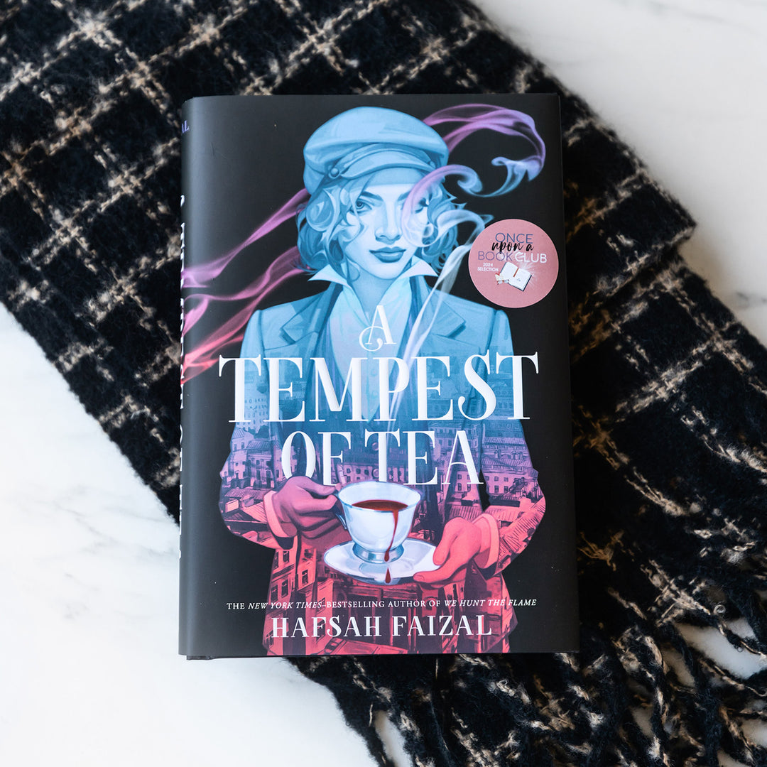 A hardcover copy of "A Tempest of Tea" by Hafsah Faizal sits on a plaid scarf lain on a white background.
