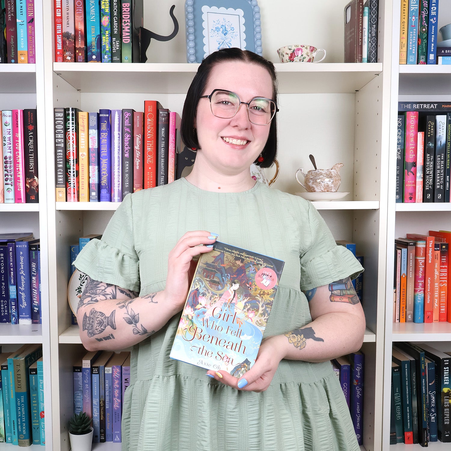 A smiling woman with short black hair, glasses, and tattoos stands in front of white bookshelves, holding a book titled 