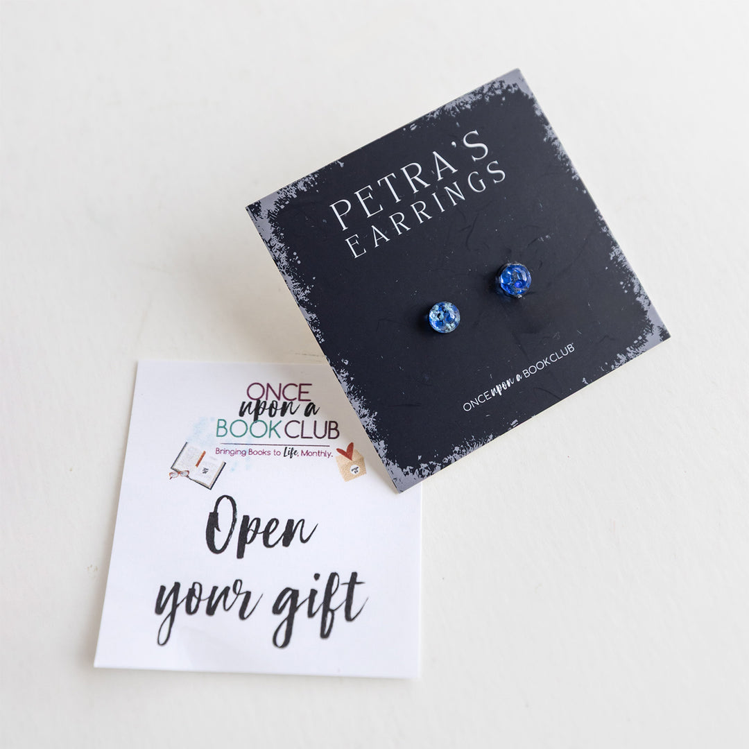 A black card labeled Petra's Earrings with two blue sapphire stud earrings next to an Open Your Gift sticky note.