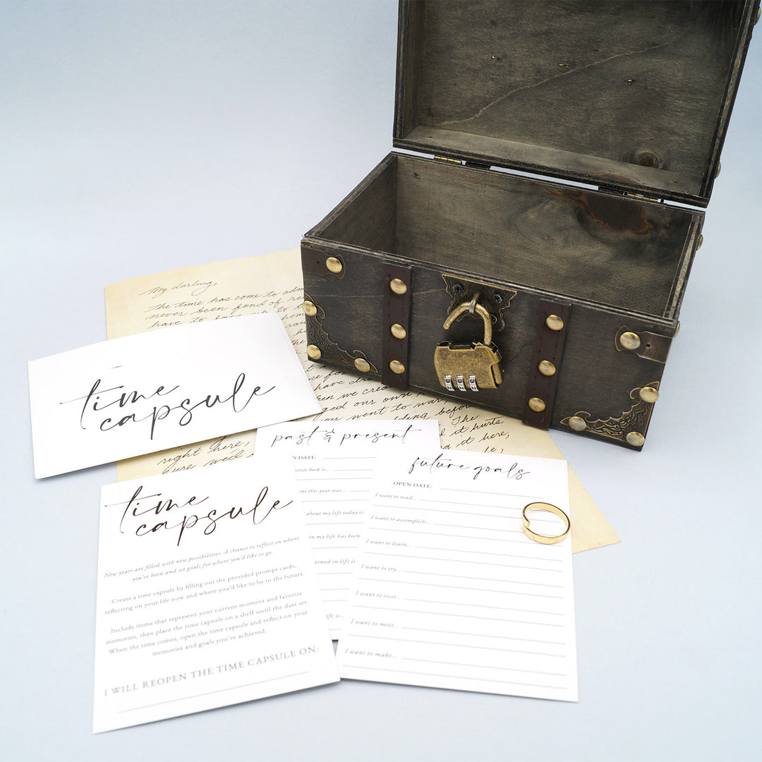a dark gray time capsule treasure chest sits open with a gold lock at the front with multiple pieces of lined paper labeled time capsule in front of it