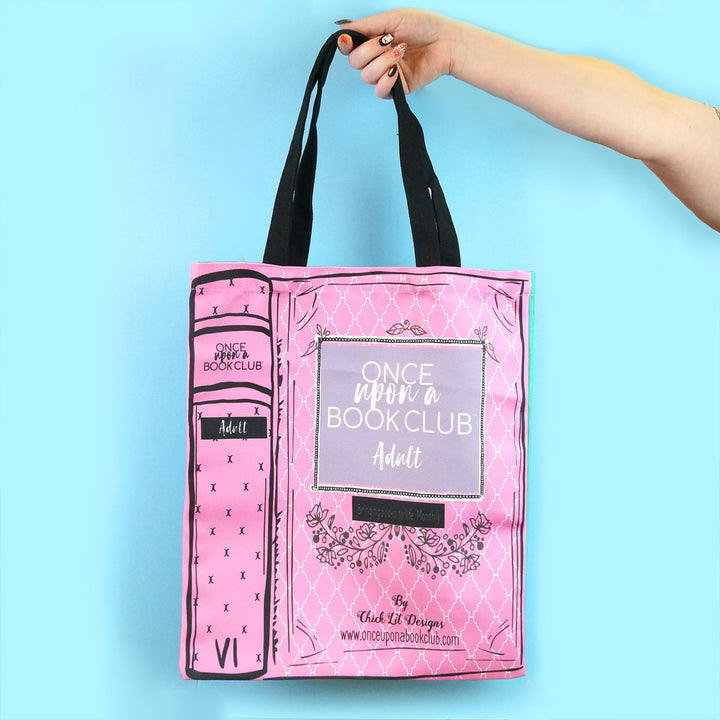 A hand holding a pink tote bag with a book-like design featuring the Once Upon a Book Club logo and the word "Adult" on the cover.