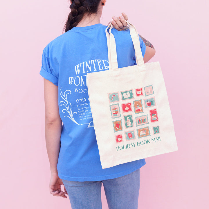 Person holding a tote bag with 'Holiday Book Mail' stamp design against a pink background