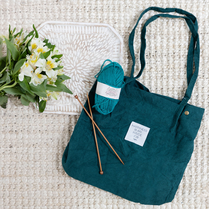 a ball of teal yarn and knitting needles sits on a dark green corduroy tote. to the left of the tote bag are white flowers