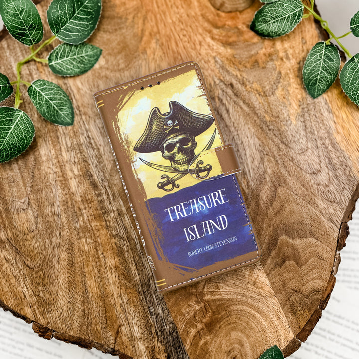 Book-shaped phone case titled 'Treasure Island' with a pirate design on a wooden surface with leaves.