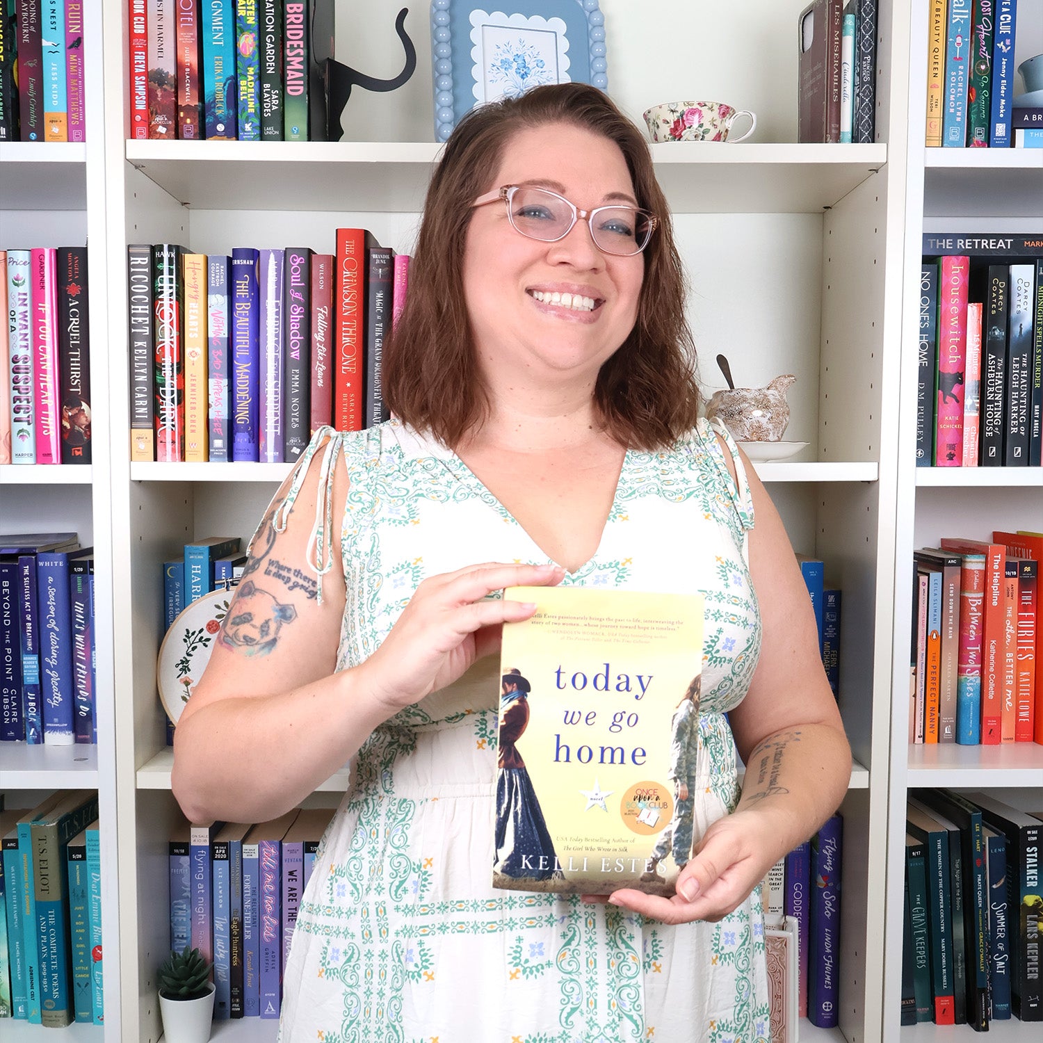 A smiling woman with medium brown hair, glasses, and tattoos stands in front of a bookshelf, holding a copy of the book 'Today We Go Home' by Kelli Estes. She is wearing a white dress with a green floral pattern.