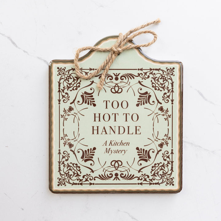 A ceramic trivet designed to look like a book cover including dark brown filigree and the words "Too Hot to Handle A Kitchen Mystery".