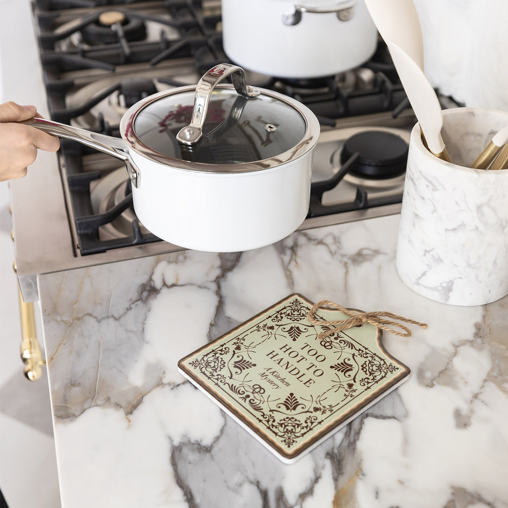 A white pot is being placed onto a ceramic trivet designed to look like a book cover including dark brown filigree and the words "Too Hot to Handle A Kitchen Mystery". The trivet sits on a marble countertop next to the stove.