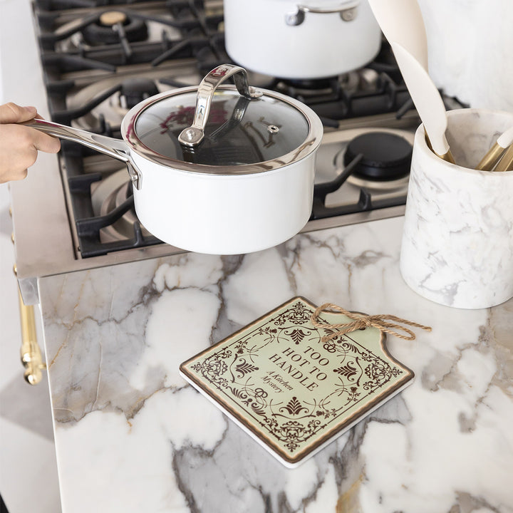 A white pot is being placed onto a ceramic trivet designed to look like a book cover including dark brown filigree and the words "Too Hot to Handle A Kitchen Mystery". The trivet sits on a marble countertop next to the stove.