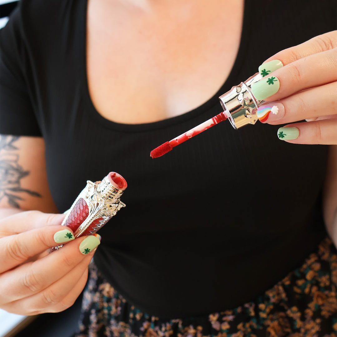 A woman with St. Patrick's Day themed nails holds a red lipstick tube with the wand out, showing a red color.