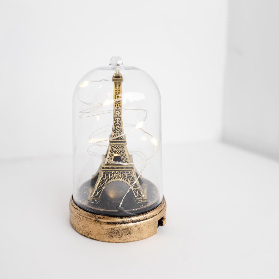 A bronze Eiffel Tower that has the words Paris France engraved in the tower design sits surrounded by small fairy lights in a glass container with a bronze base.
