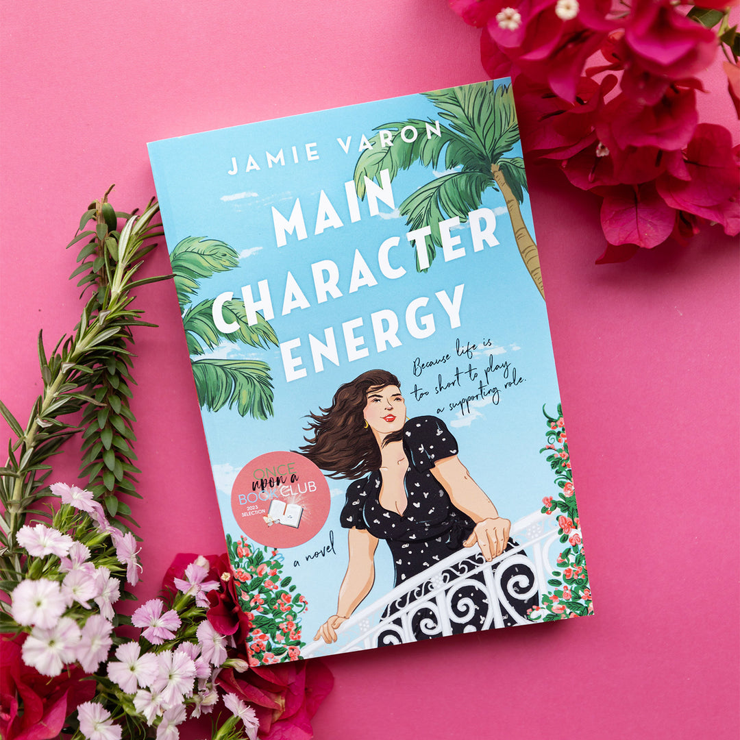 paperback book "Main Character Energy" by Jamie Varon on bright pink background with pink and white flowers