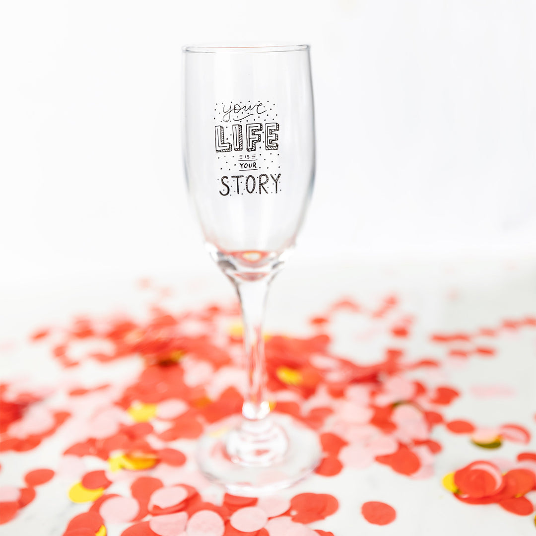A champagne flute with the phrase "your LIFE your STORY" printed on it, placed on a surface scattered with red, pink, and gold confetti.