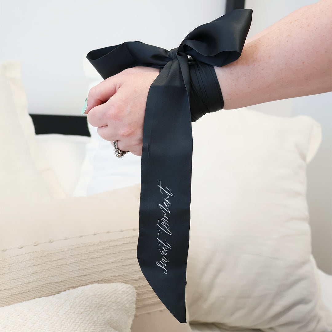 Two hands are tied at the wrist with a satin black tie. On the tie in white cursive it says Sweet Torment.