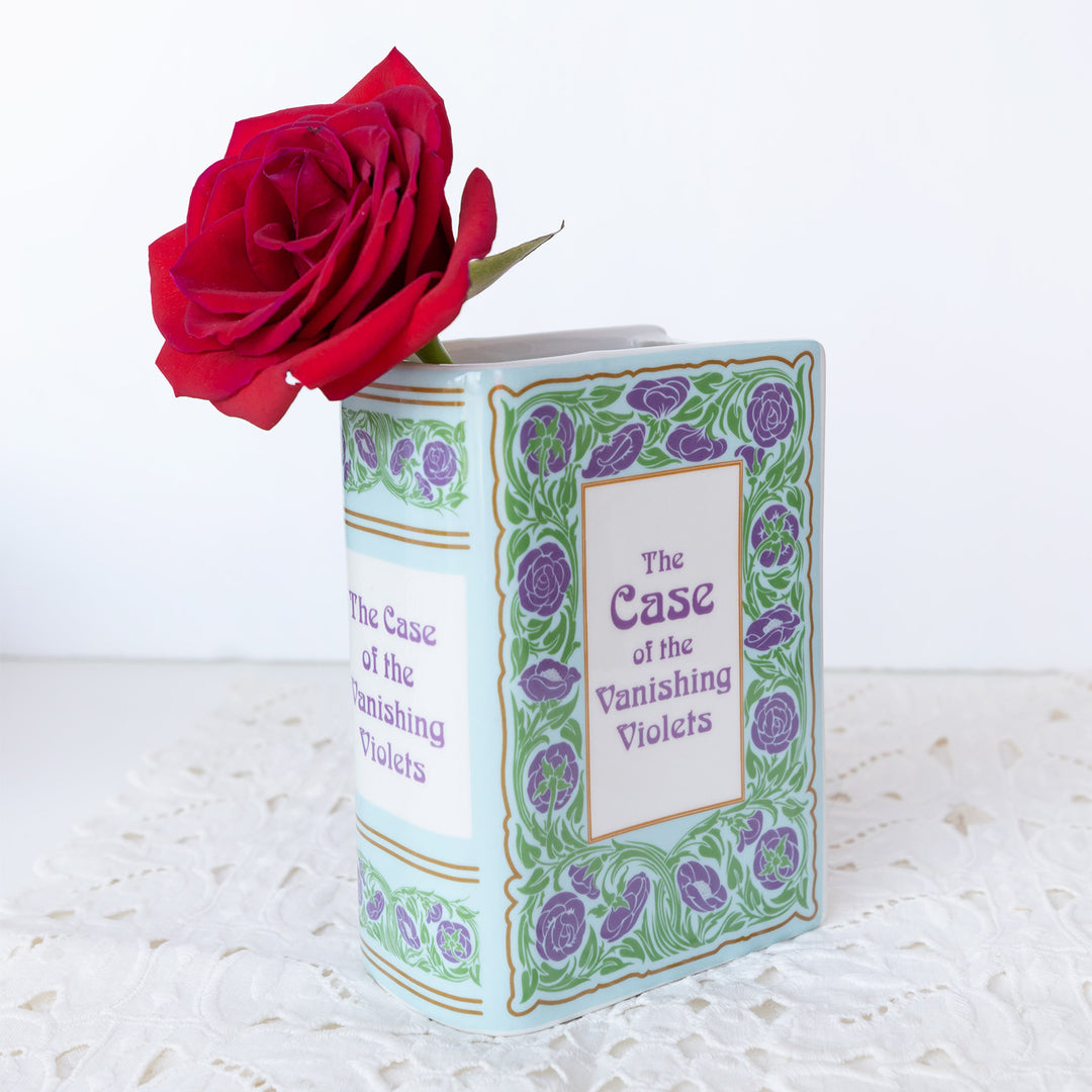 Book shaped vase labeled The Case of the Vanishing Violets in green and purple florals with a red rose sticking out of the top