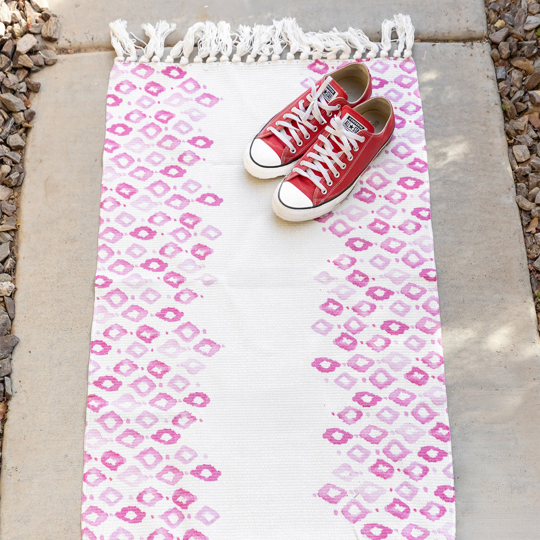 A pair of red converse shoes lay on a tasseled door mat outside on the sidewalk. The door mat is white with pink diamond pattern on the left and right sides with the center blank.