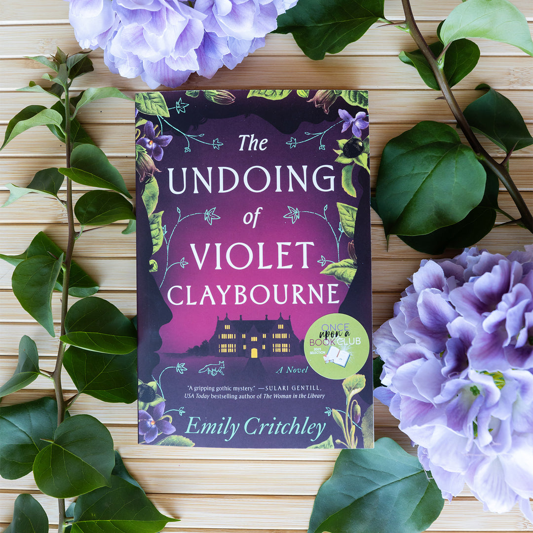 Book titled 'The Undoing of Violet Claybourne' by Emily Critchley surrounded by purple flowers and green leaves on a wooden surface.