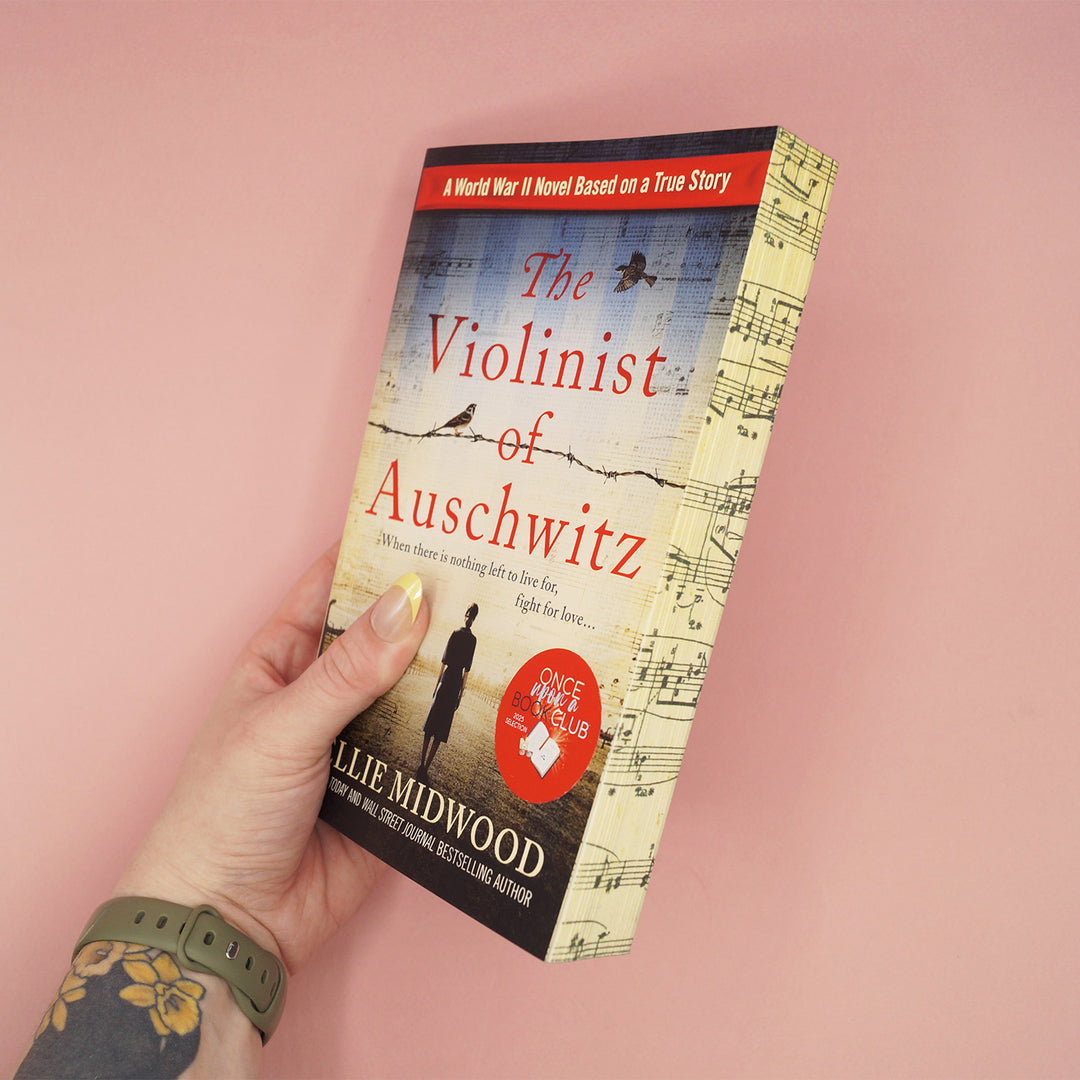 Book titled 'The Violinist of Auschwitz' by Ellie Midwood showcasing patterned page edges is held by a hand on a pink background