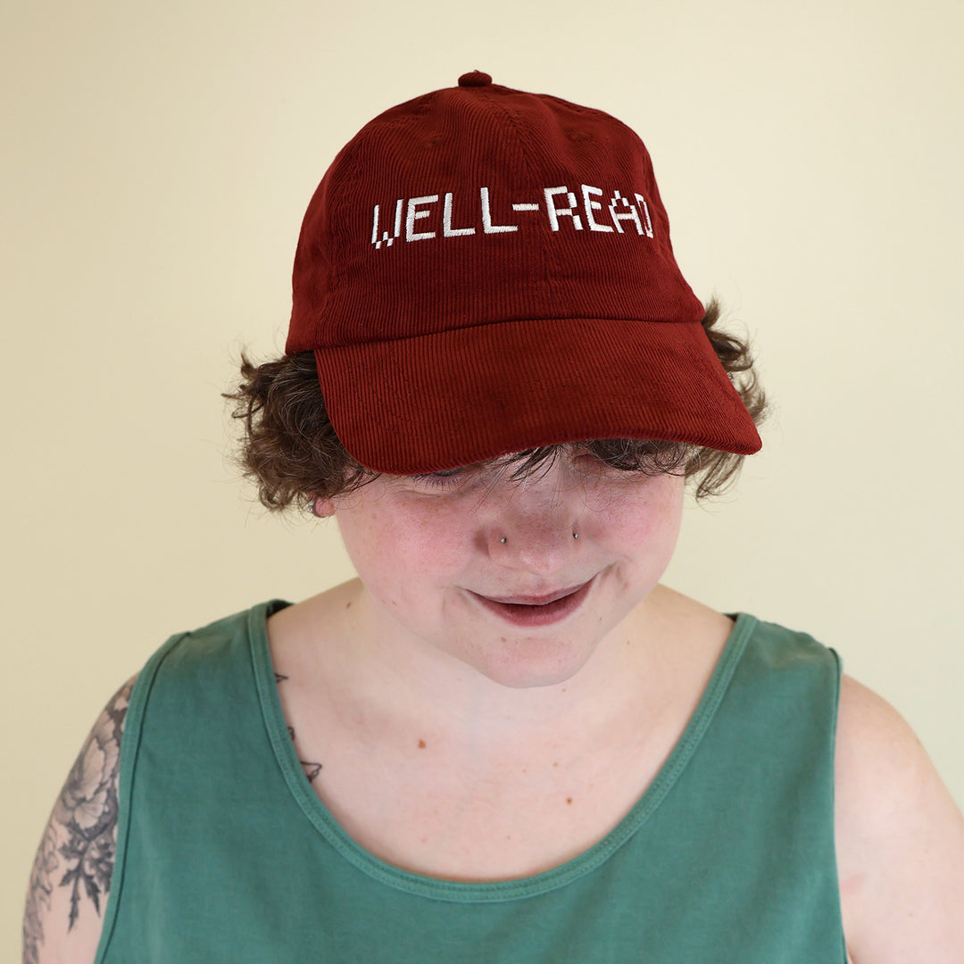 Woman wearing a maroon corduroy cap embroidered with 'WELL-READ' text on a beige background