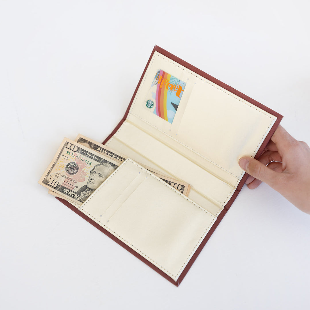 A white hand holds a red-brown wallet open, showing a beige interior with a gift card and cash tucked into the folds.