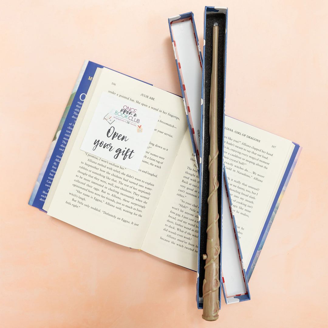 An open book with an Open Your Gift sticky note lays flat on a pink background. Inside an open box on top of the book lies a magic wand.