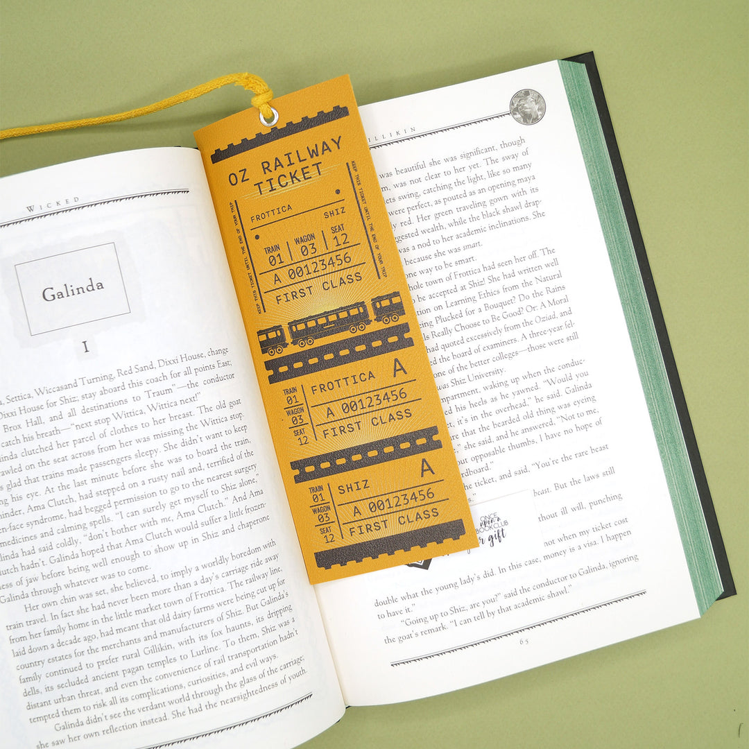 A gold train ticket bookmark with a gold tassel lays in an open book with green sprayed edges. The bookmark says Oz Railway Ticket and has information about the train ride from Frottica to Shiz.