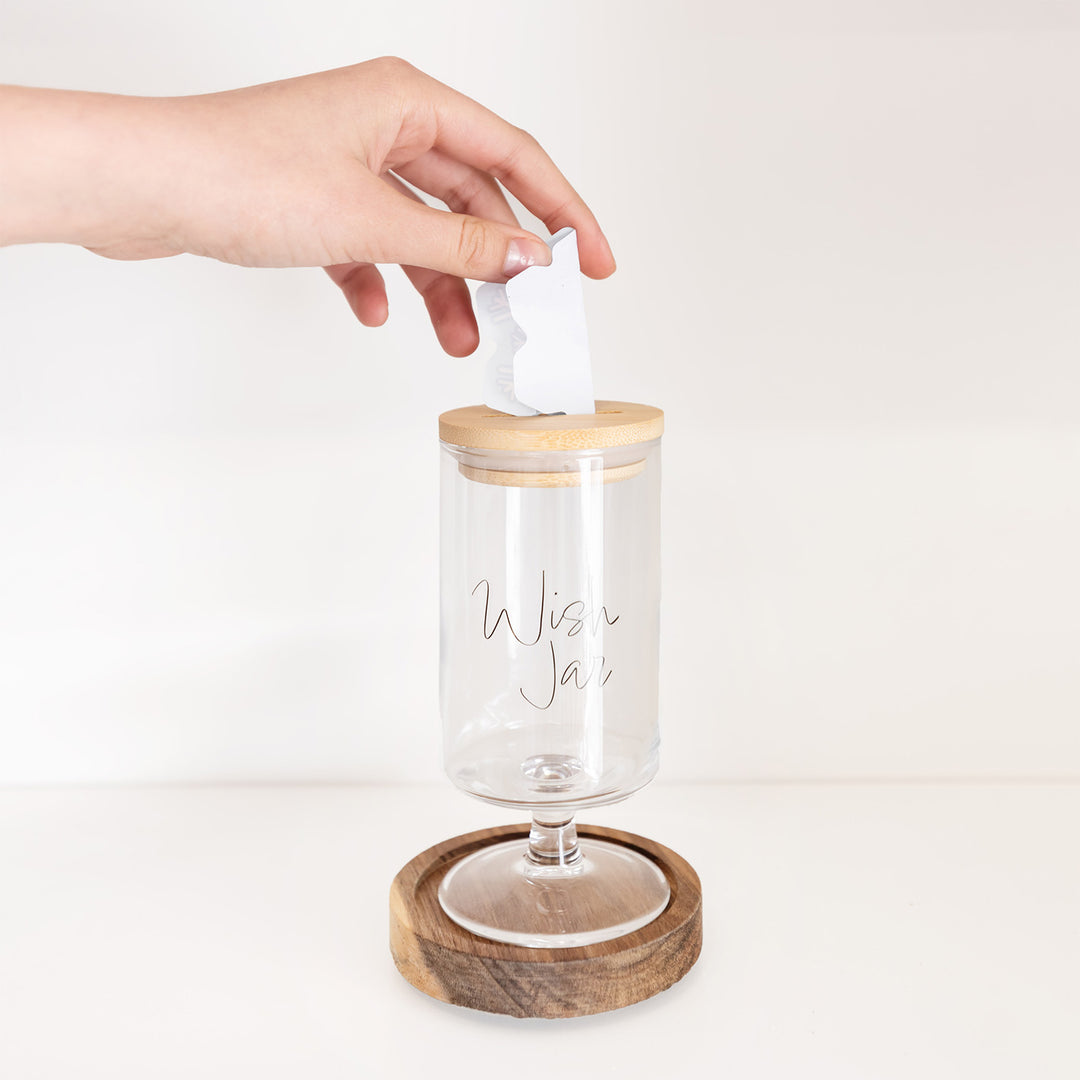 A hand places a folded piece of paper shaped like a snowflake into a glass jar labeled Wish Jar