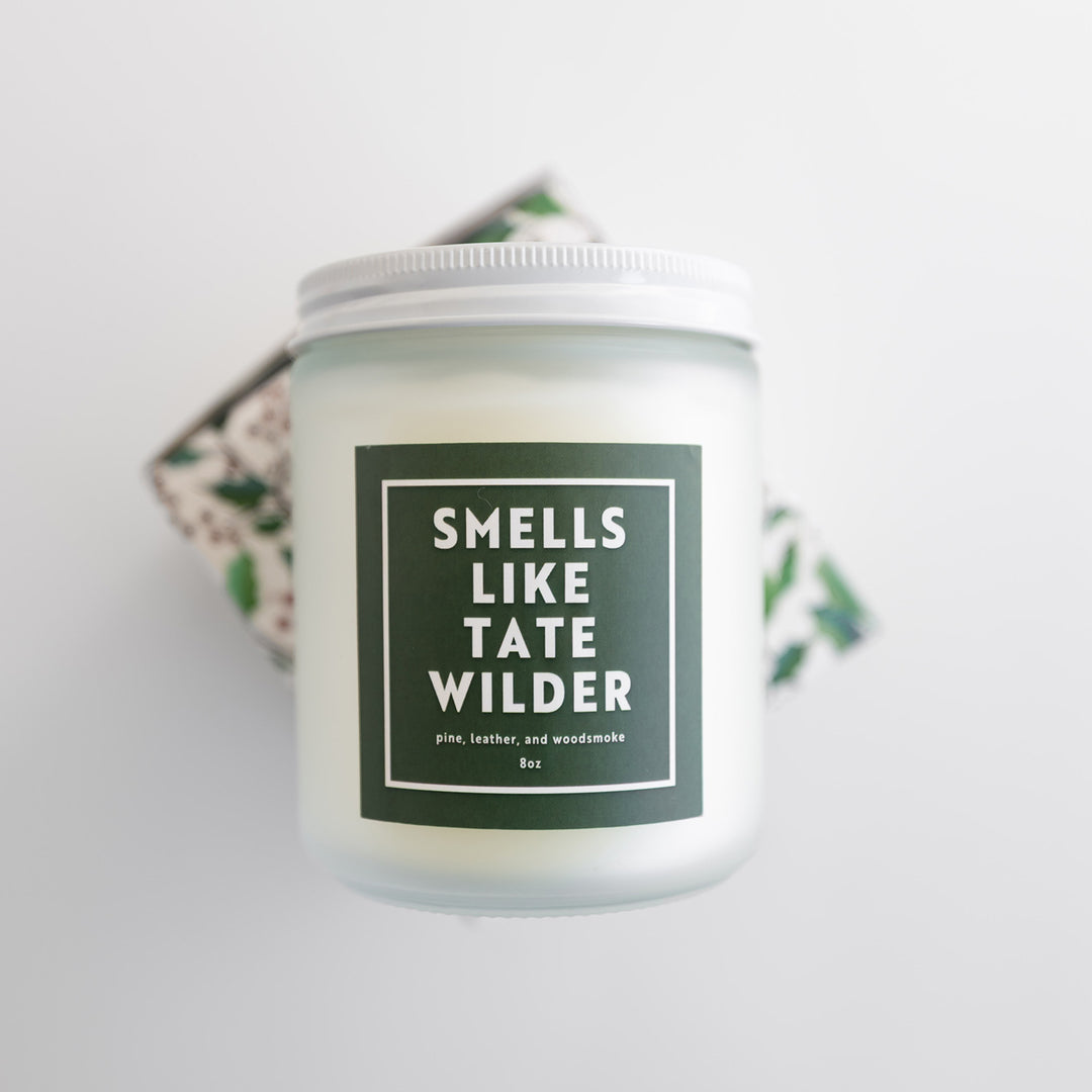 A white candle with a green square label that says Smells Like Tate Wilder. Pine, leather, and woodsmoke.