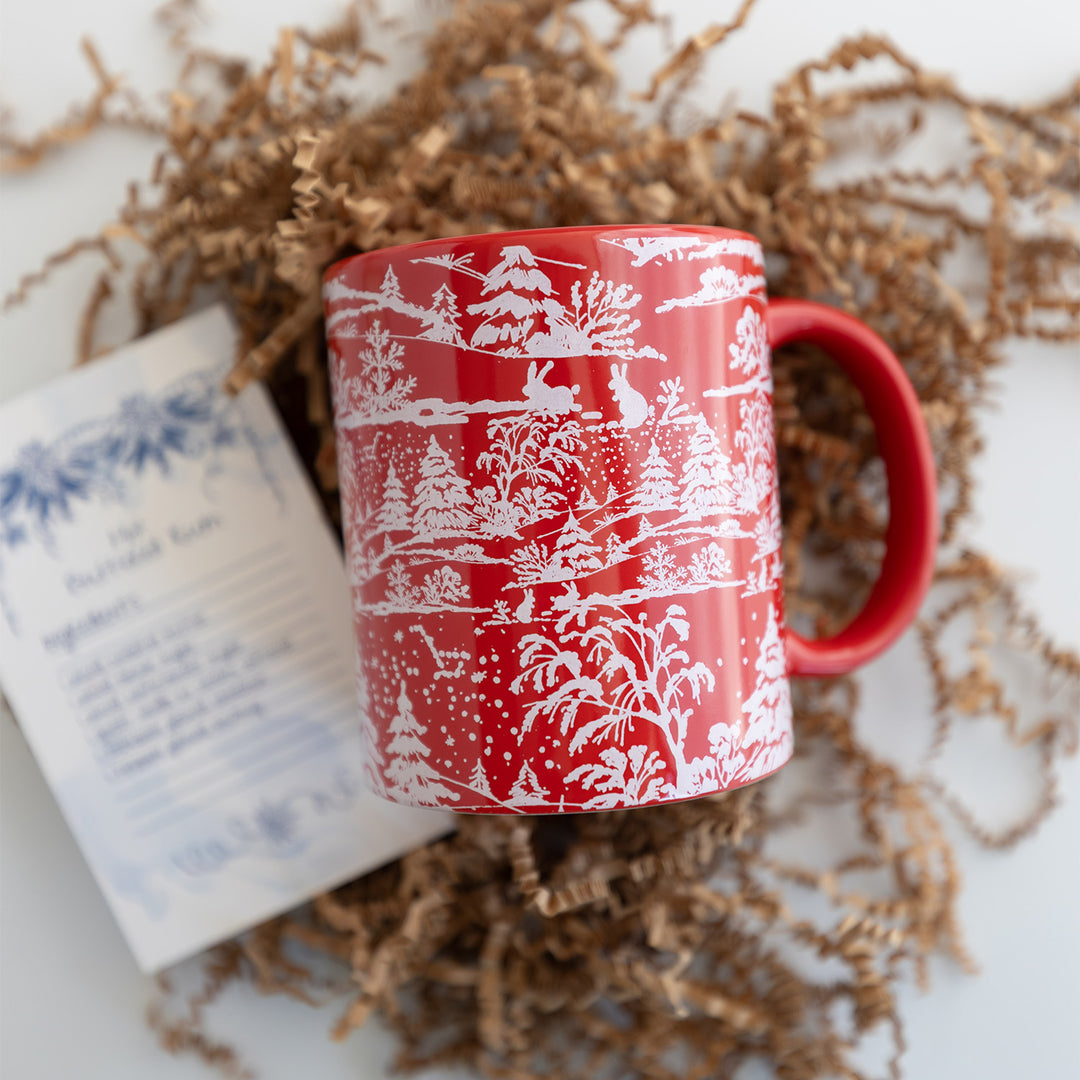 A red mug with a white winter scene on it next to a recipe card.
