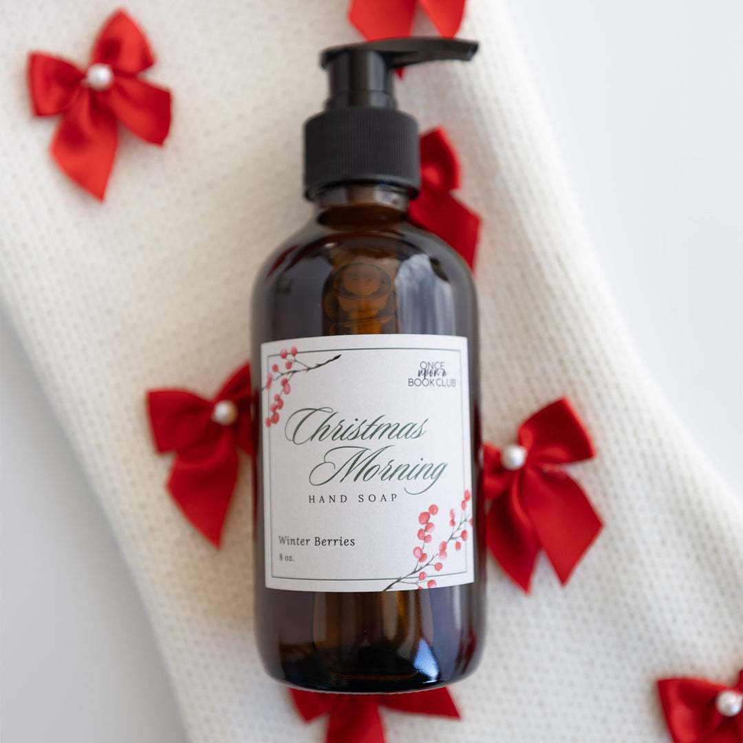 A bottle of soap with a pump dispenser in it labeled Christmas Morning Hand Soap Winter Berries. The bottle lays on a white fabric with mini red bows.