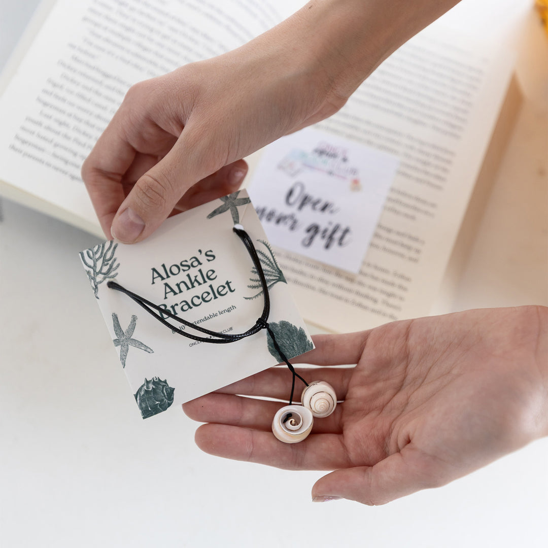 Hands hold a card labeled Alosa's Ankle Bracelet with a black thread ankle bracelet  with two seashells on it attached. In the background is an open book with an Open Your Gift sticky note.