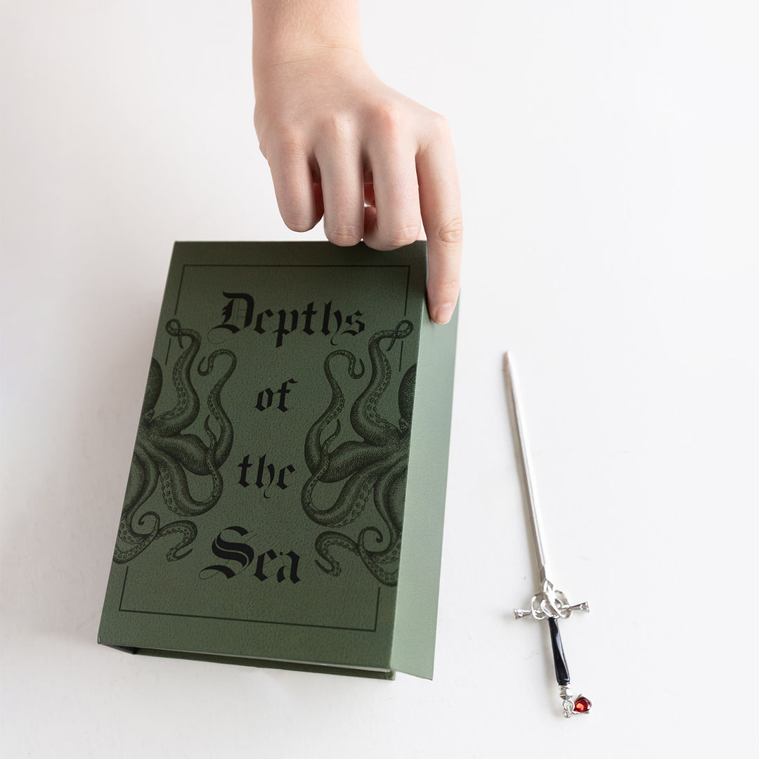 A hand opens a green book-shaped storage box. The front of the box features the text Depths of the Sea and two drawings of an octopus. There is a mini dagger next to it with a red gem attached to the handle.