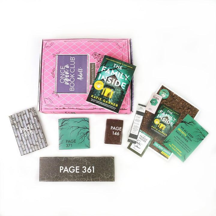Paperback edition of The Family Inside lays on a pink OUABC box. In front of the box are four themed wrapped gifts labeled with page numbers and paper items.