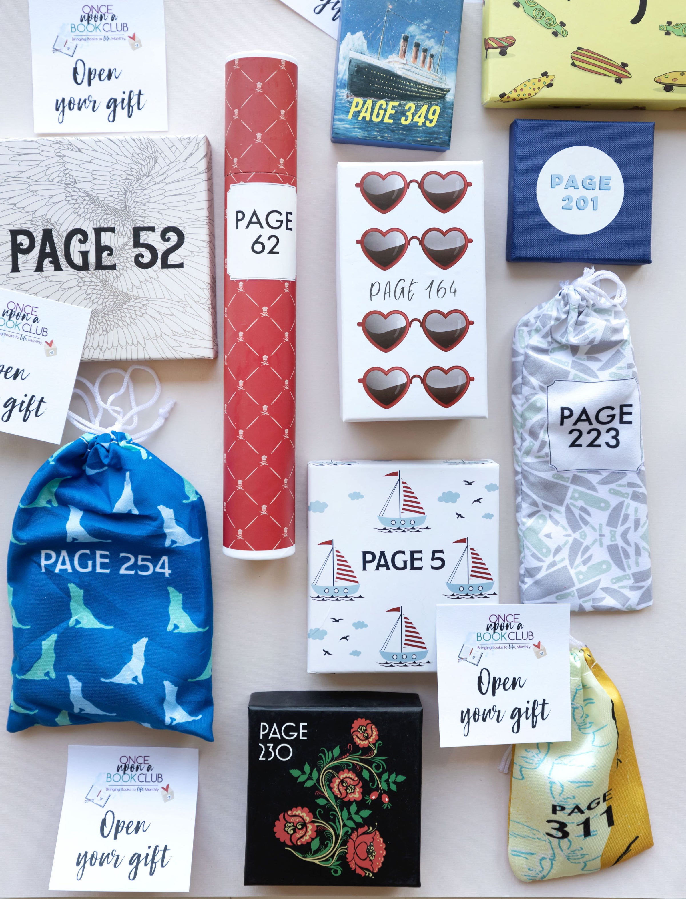 A collection of wrapped gifts labeled with page numbers lay flat on a surface. Gift packaging varies from boxes, to bags, to tubes. Colors vary but all gifts are labeled with a page number. There are also some Open Your Gift stickers around the image.
