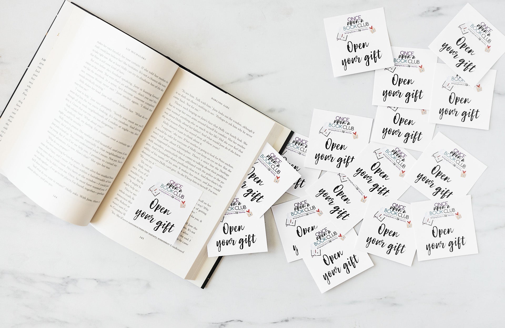 An open book lays on a marble counter with a pile of "open your gift" sticky notes coming out of the side and reaching all the way to the top right of the image.