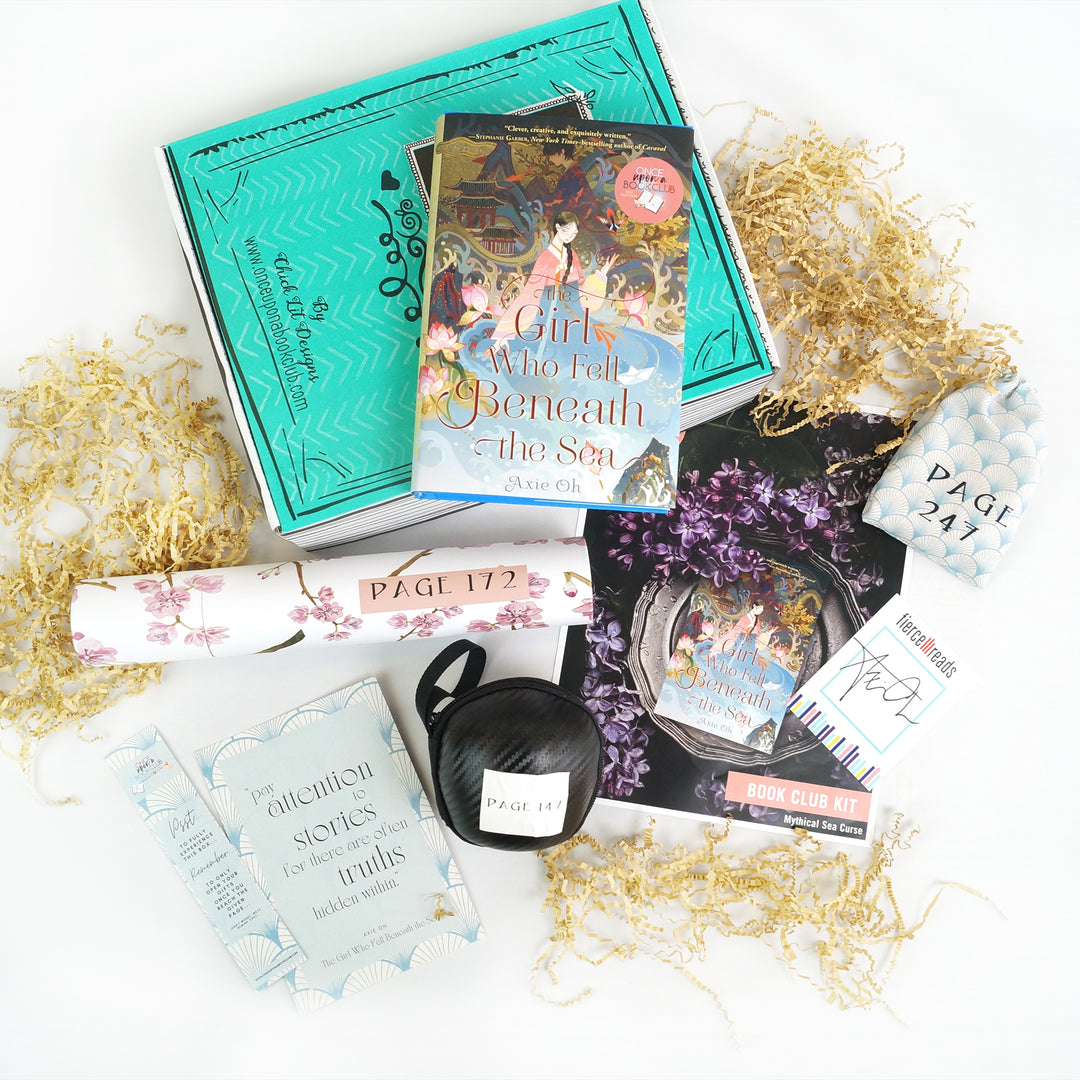 A hardcover edition of The Girl Who Fell Beneath the Sea by Axie Oh lays on a green Once Upon a Book Club box. In front of the box are a white tube, bookmark, quote card, black ball, bookclub kit, signature card, and white drawstring bag. The bag, tube, and ball are all labeled with page numbers.