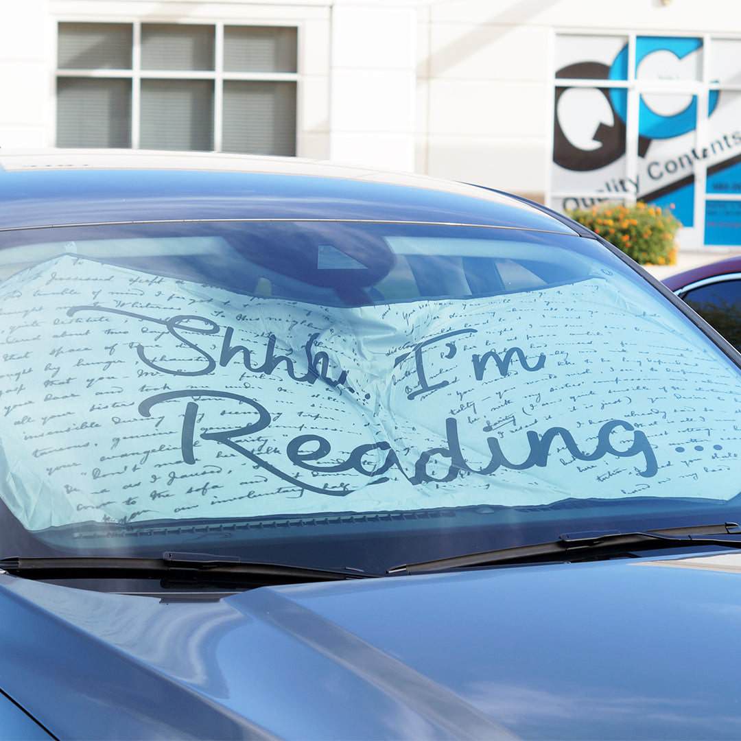 A white sunshade is in the front windshield of a car. The sunshade has black script pattern across it. The center in bold black script says "Shhh, I'm Reading"