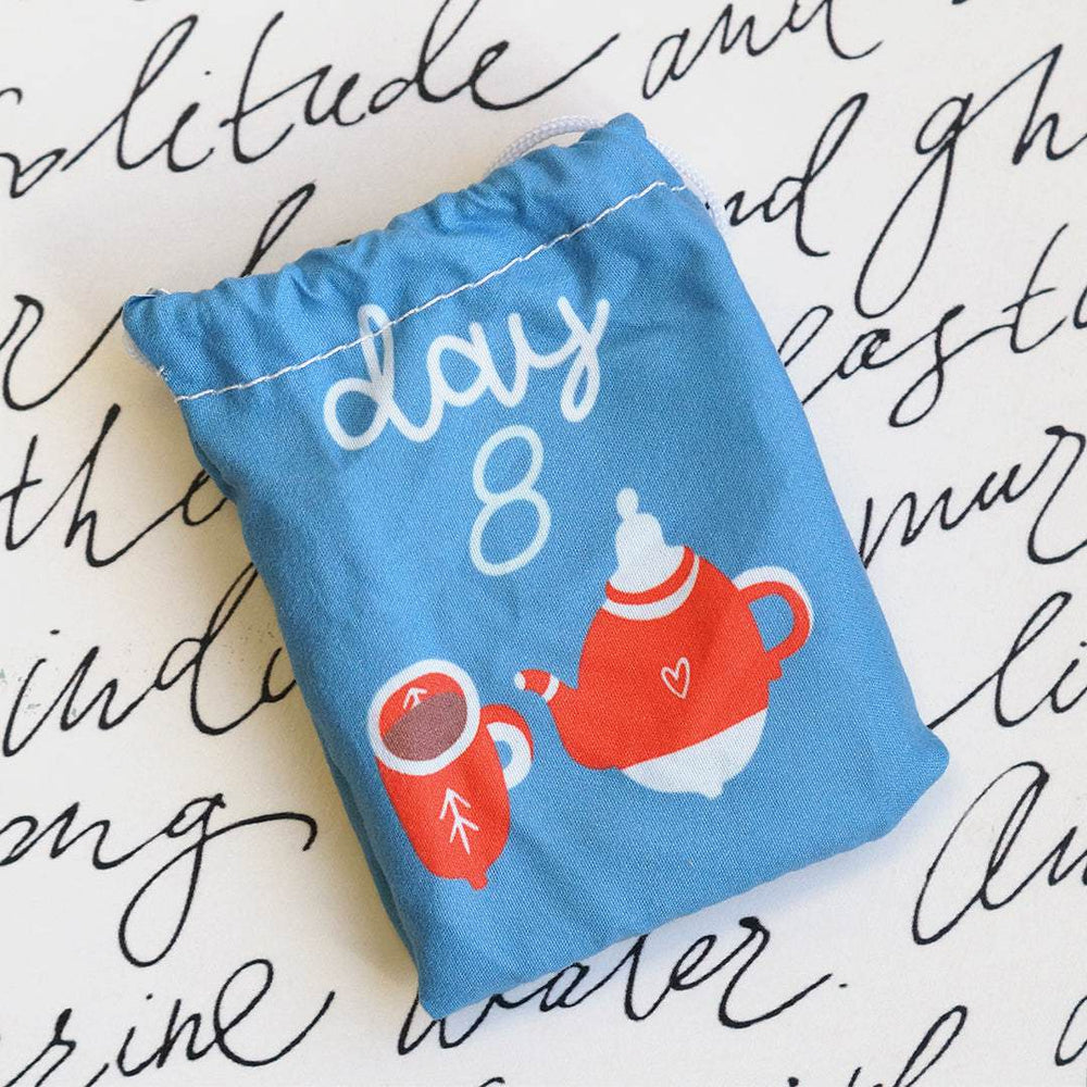 a blue drawstring bag with an image of a mug and teapot with the words "Day 8" lays on a white background with black script