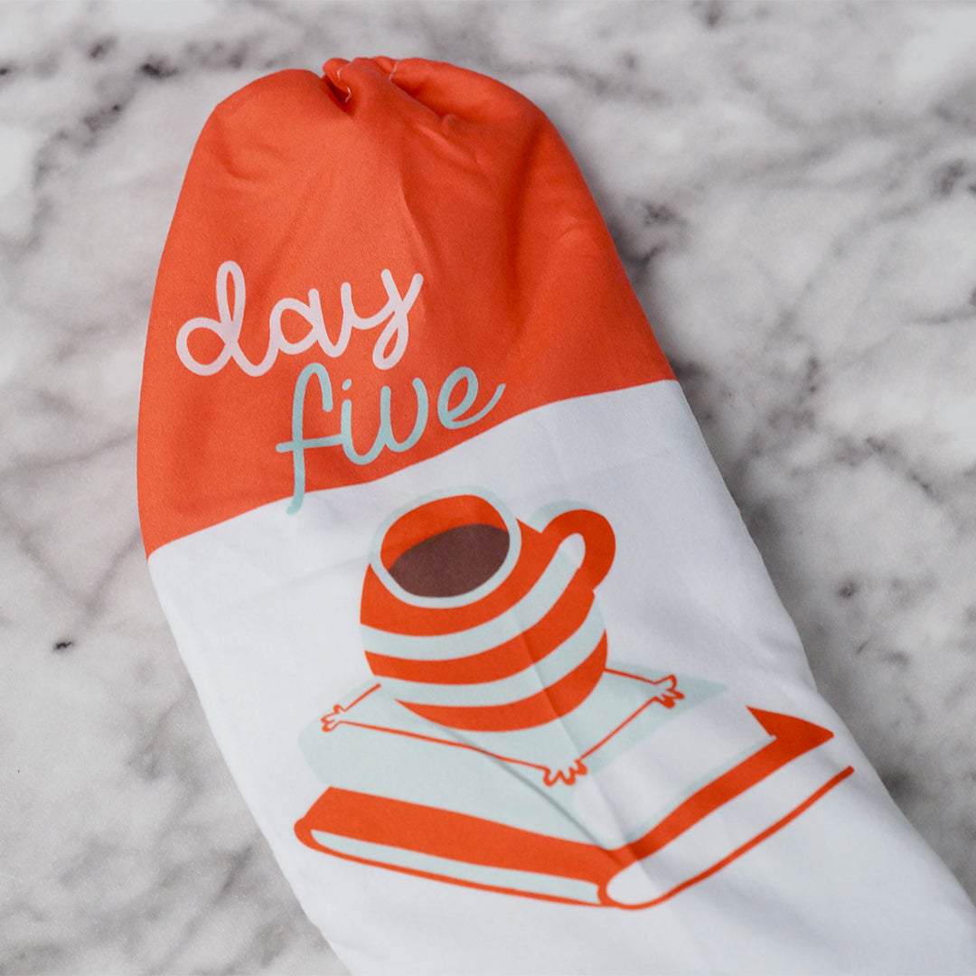 a orange and white drawstring bag labeled Day Five with a coffee cup on a stack of two books