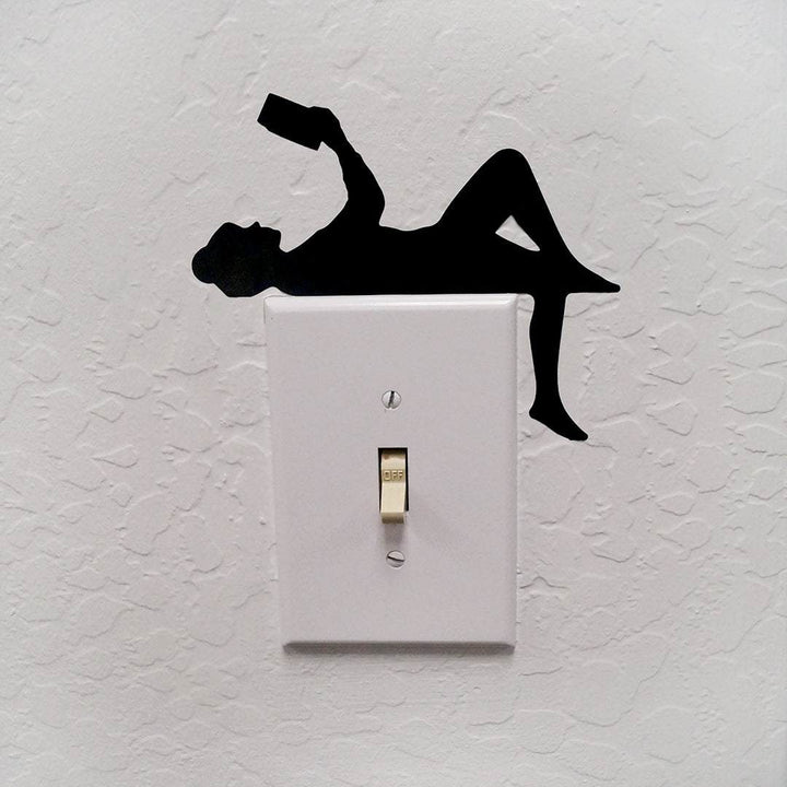 A light switch decal on top of a light switch. The decal is a black silhouette of a girl laying down reading, holding a book above her face