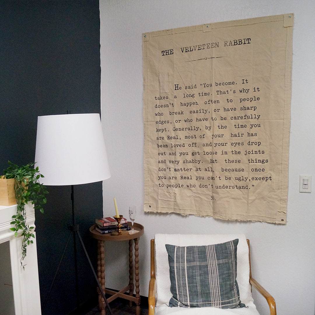 a canvas wall tarp labeled The Velveteen Rabbit with a quote below it hangs on the wall above a chair and lamp
