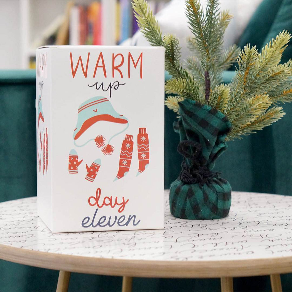 A white box with a photo of a hat, gloves, and socks labeled "Warm Up Day Eleven" sits on a white table next to a plant