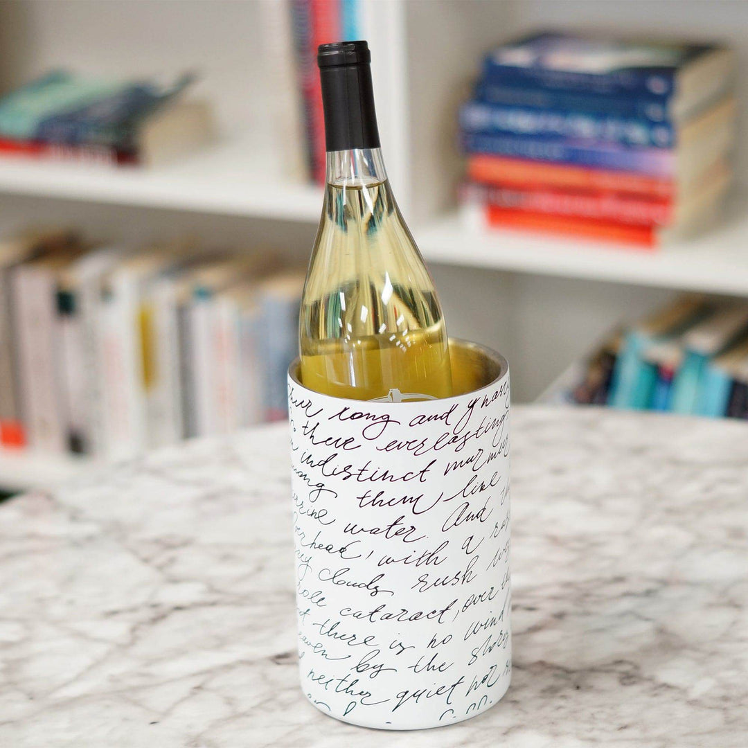 A white wine chiller with black script across it holding a bottle of white wine. The wine chiller is sitting on a table in front of a book shelf.