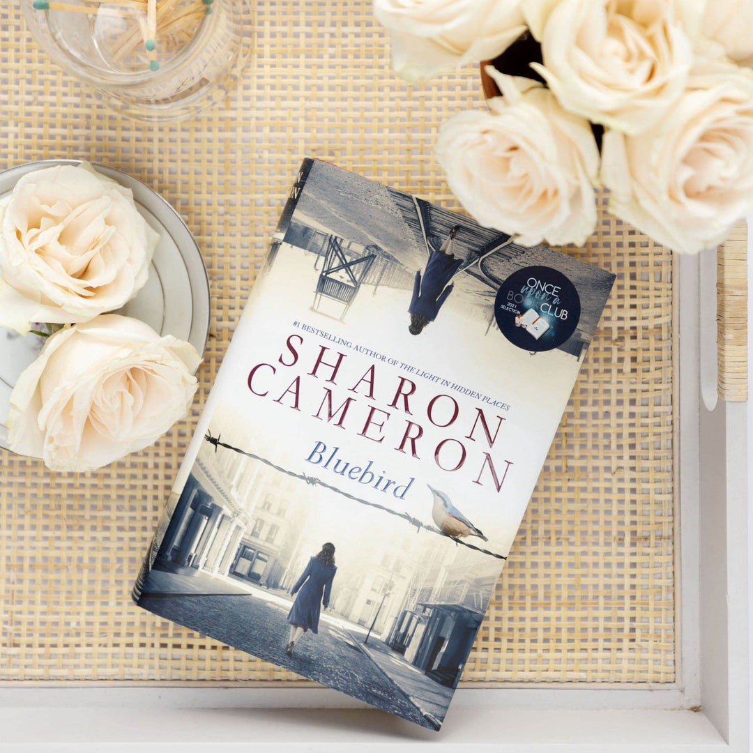 a hardcover edition of Bluebird by Sharon Cameron lays next to white roses